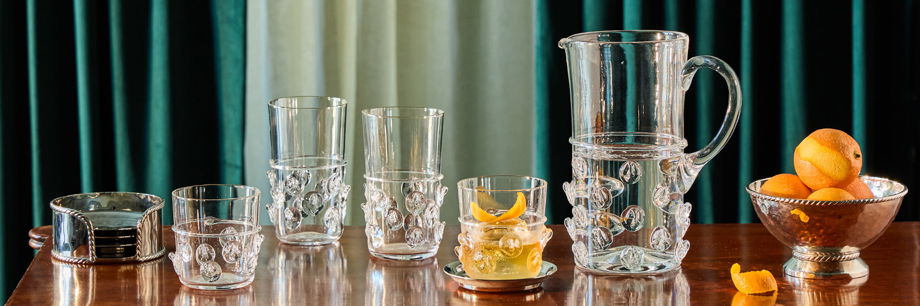 Set of glass tumblers and a pitcher on a wooden table with oranges and graham cocktail bowl.