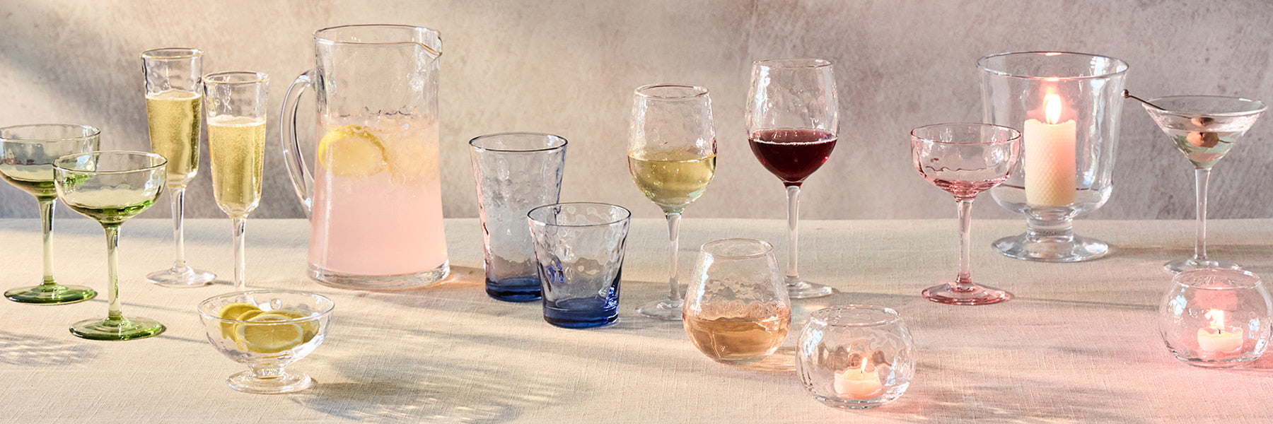 Glassware including a pitcher, wine glasses, and cocktail glasses on a linen tablecloth. The glassware featured comes in a variety of colors such as green, pink, blue and clear.