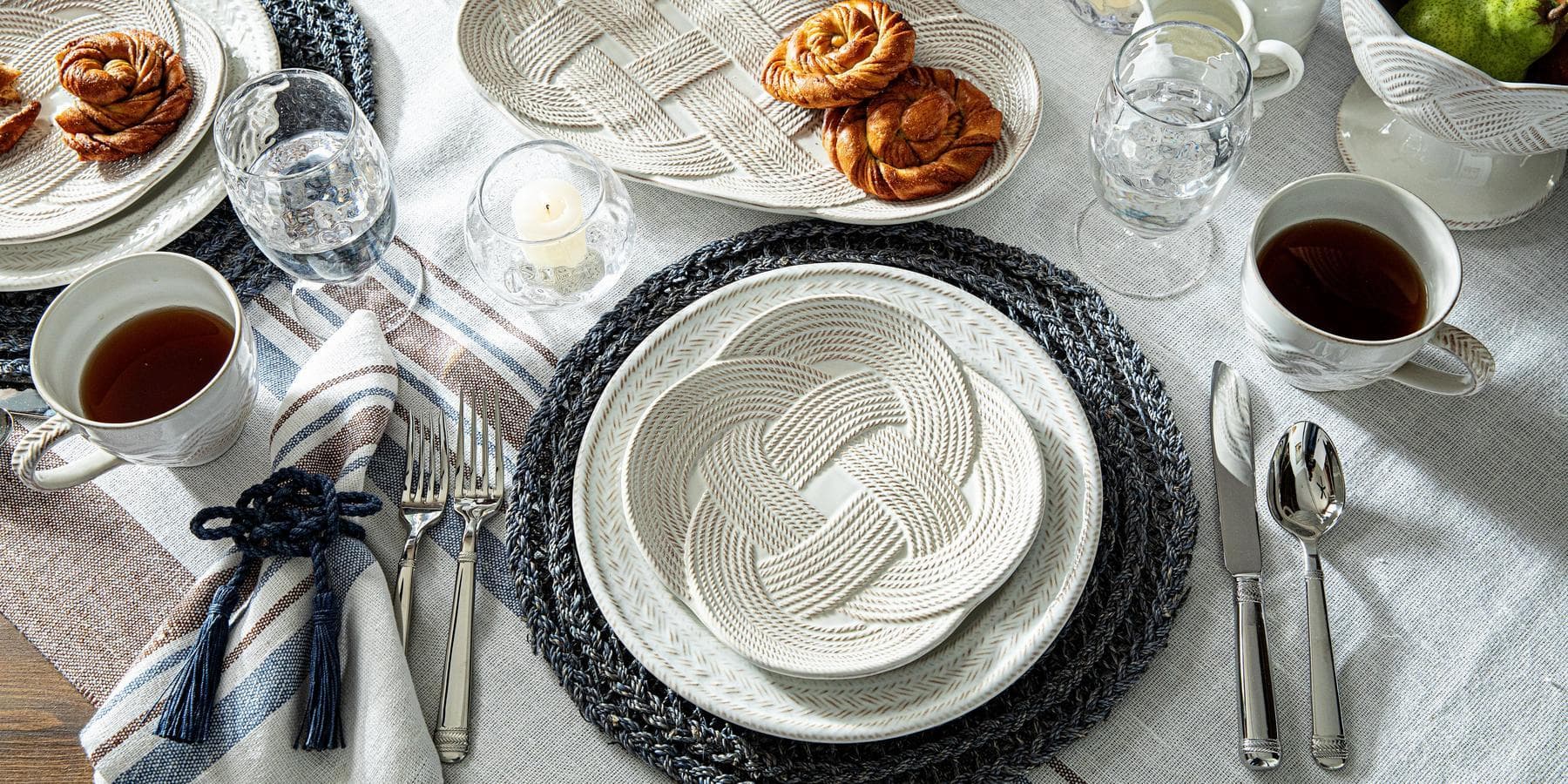 Dining table setting with woven plates, cutlery, and tea cups on a white tablecloth.