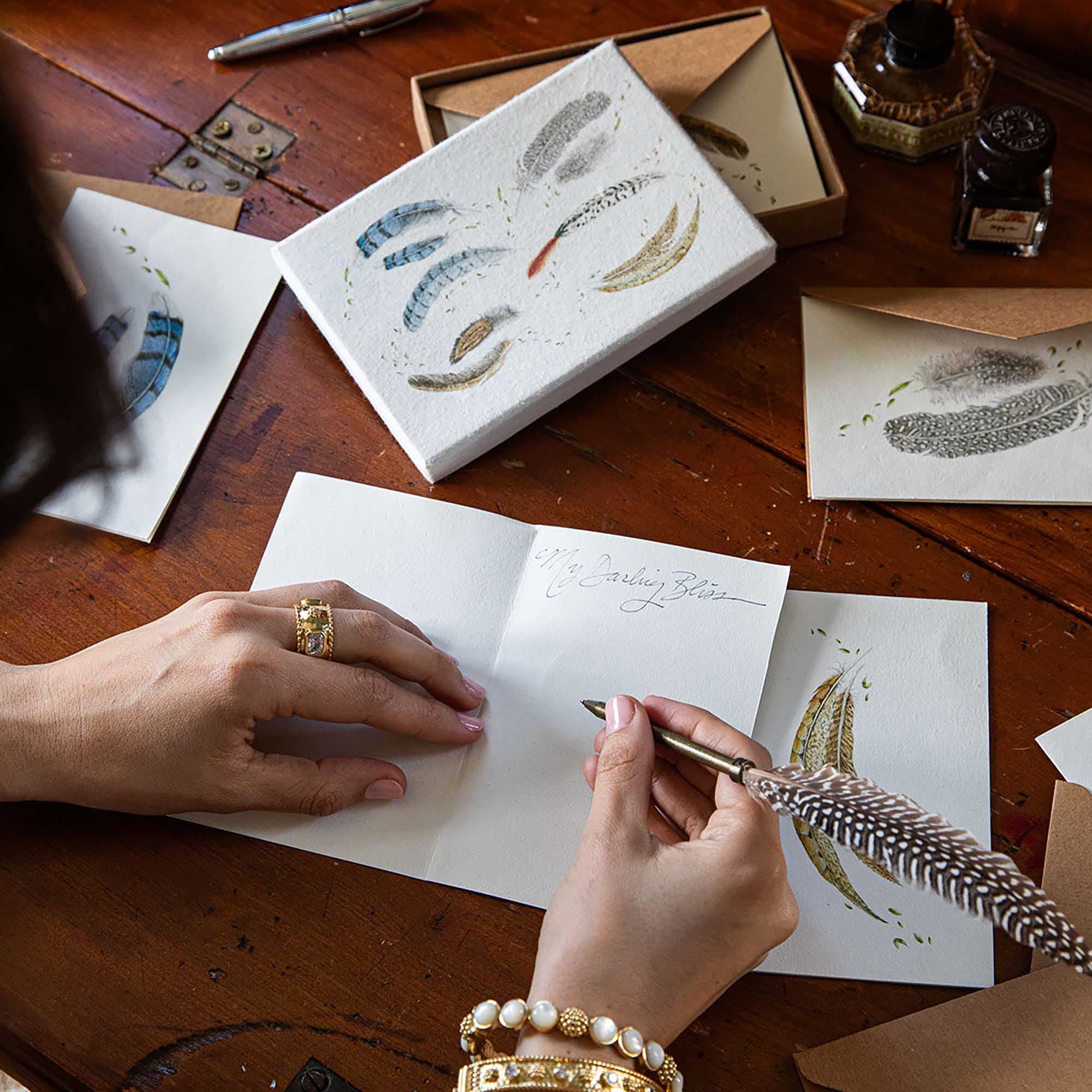Person writing with a feather pen on a wooden desk with stationery items.