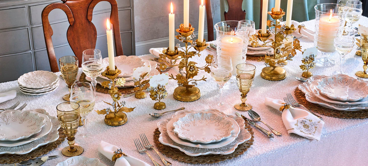 An elegant table setting with white plates, gold-toned goblets, lit taper candles in ornate gold holders, and a white tablecloth in a dining room setting.