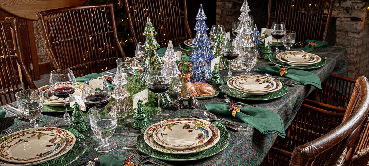 A festive holiday table setting with layered green plates, dark green napkins, wine glasses, and decorative glass and metal Christmas trees on a patterned tablecloth in a rustic dining room.