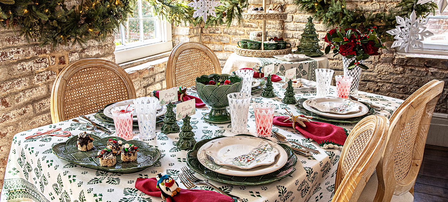 Decorated Christmas table with festive tablecloth, candles, and small trees in a cozy room.