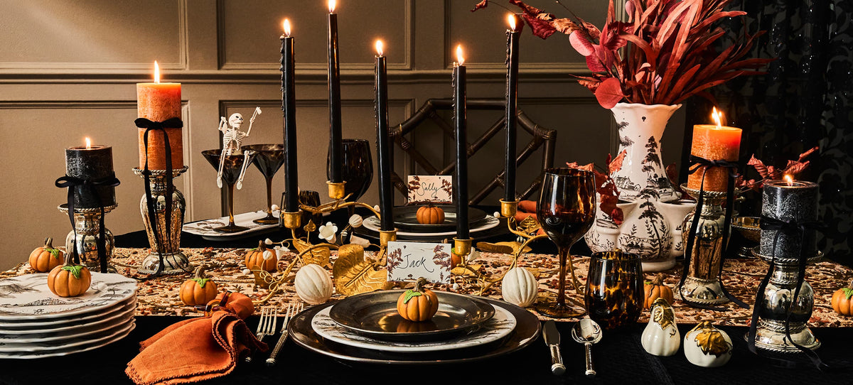 A Halloween-themed table setting with lit candles, decorative pumpkins, black candlesticks, place cards, and ornate candle holders.