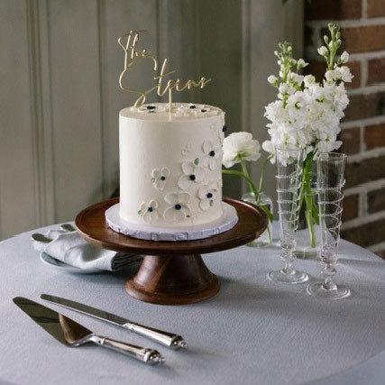White cake with decorative elements on a wooden stand, accompanied by flowers and glasses.