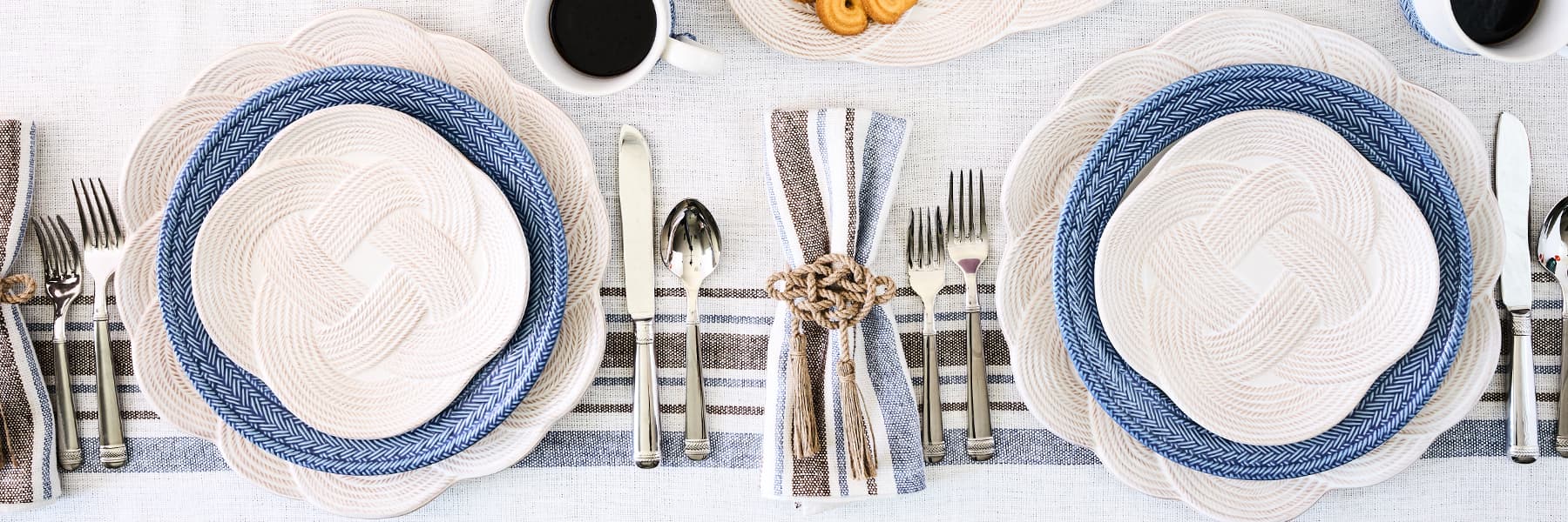 Table setting with white plates, blue rims, silverware, and striped tablecloth.