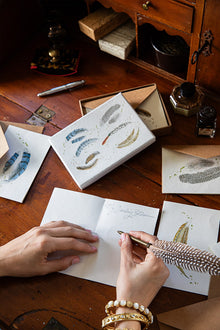 Person writing with a feather pen on a wooden desk with stationery items.