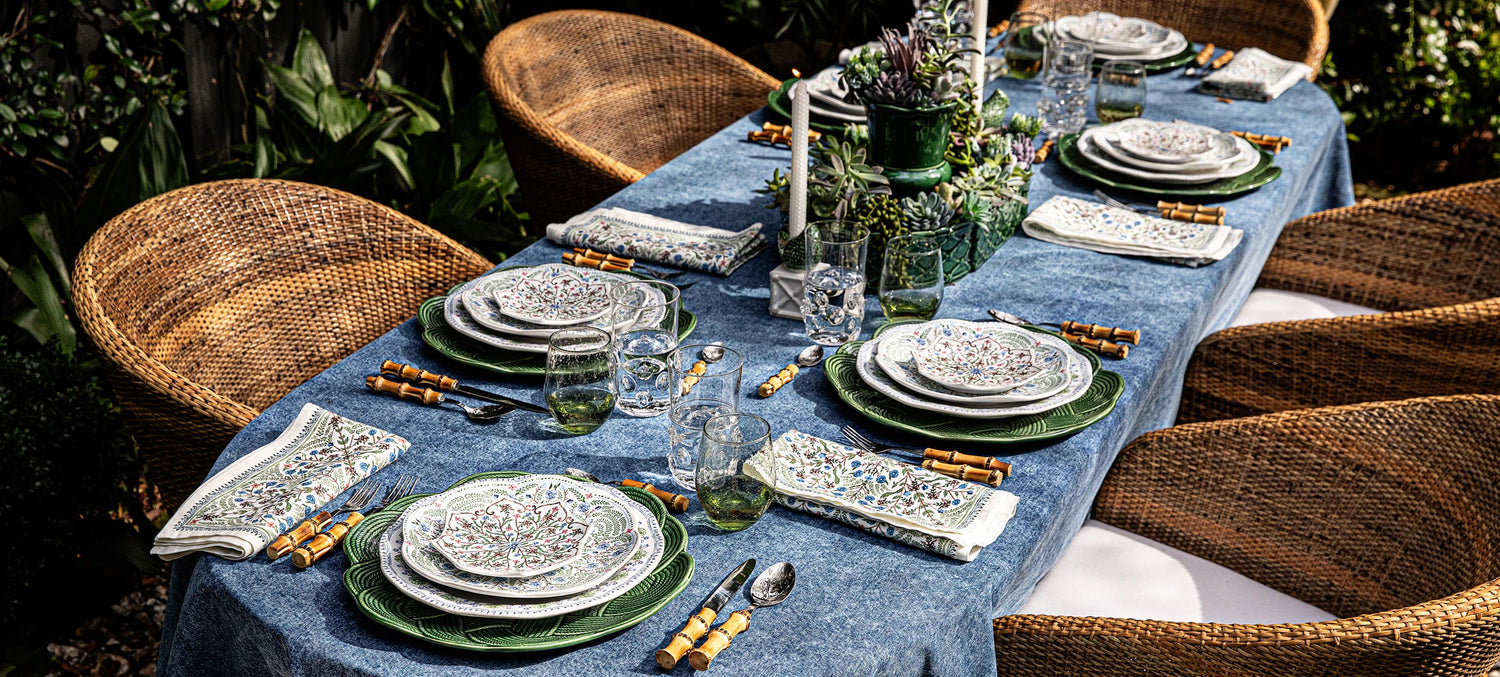 Dining table set with plates, glasses, and cutlery on a blue tablecloth outdoors.