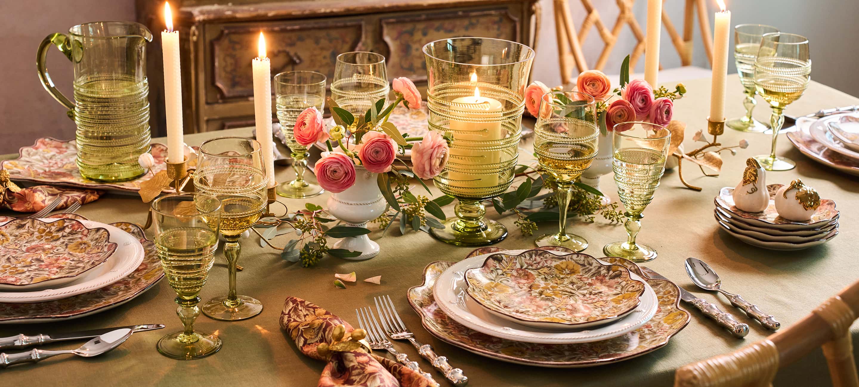 An elegantly set dining table with floral centerpieces, lit candles, clear glassware, and patterned plates in a warm, rustic indoor setting.