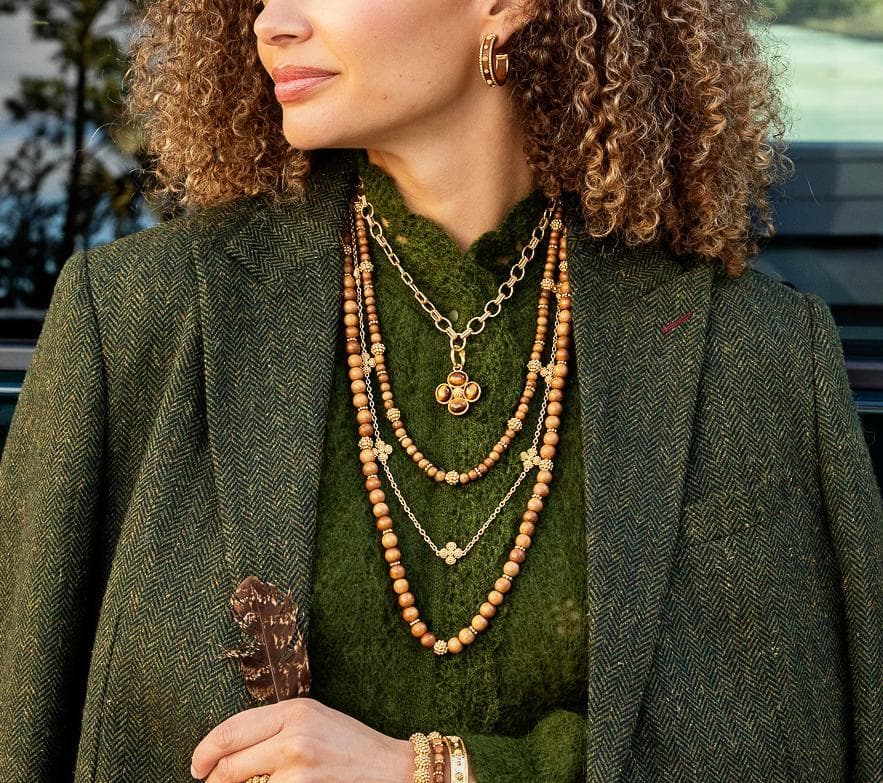 A close-up lifestyle shot of a woman wearing a layered necklace set. The set includes a gold chain with a clover pendant, a multi-strand wooden bead necklace, and a delicate silver chain with a star charm, styled with a green knit sweater and textured green blazer.
