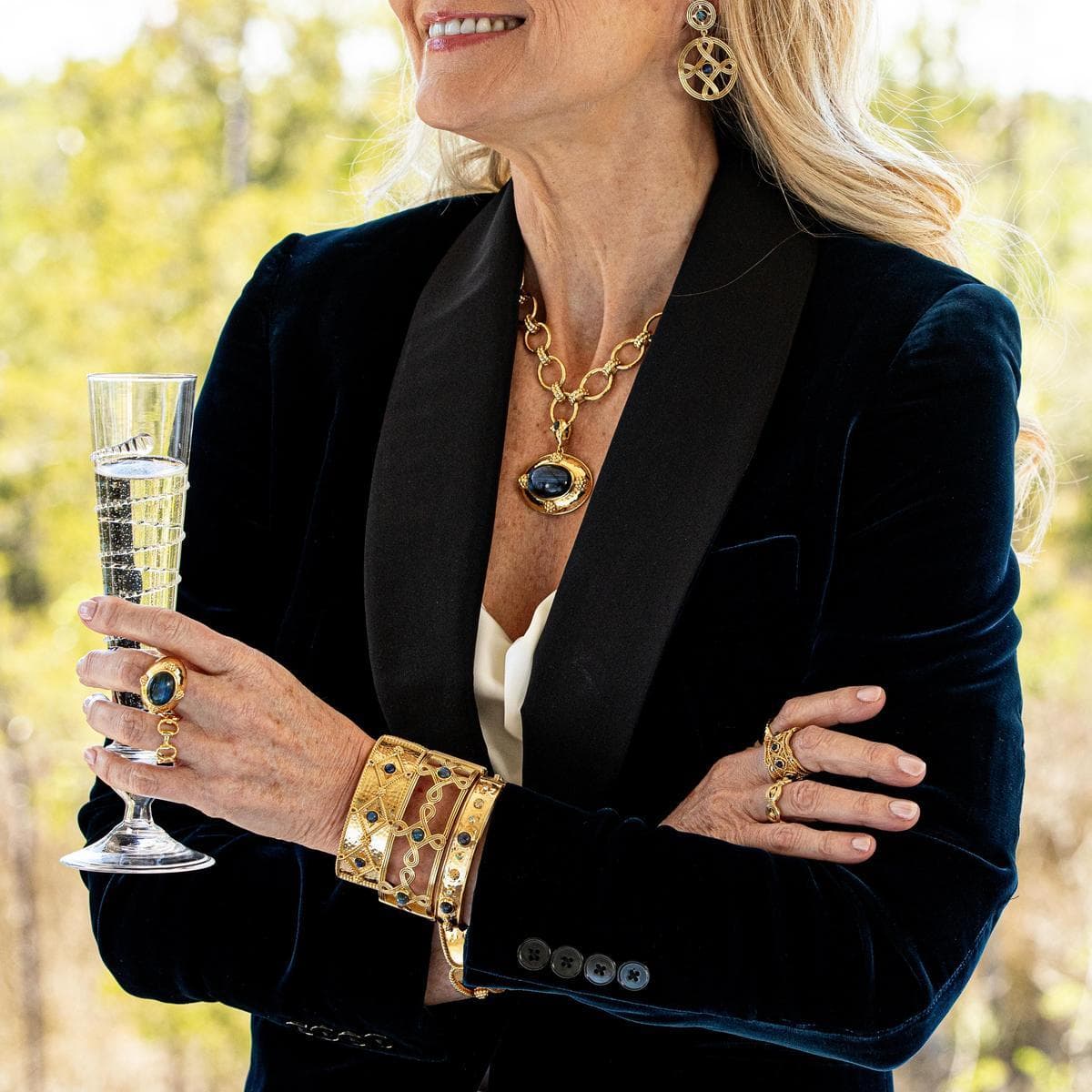 Woman in a black blazer holding a glass of champagne, wearing gold jewelry outdoors.