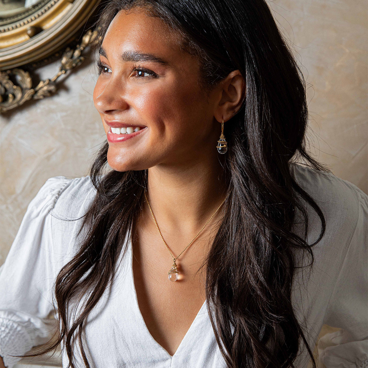 Woman wearing a white blouse with jewelry, sitting in an elegant room.