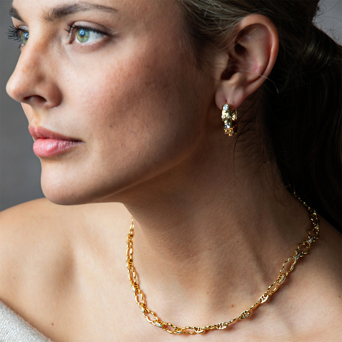 A close-up of a woman wearing a delicate gold chain necklace and a matching gold hoop earring with clear gemstones, shown against a soft gray background.