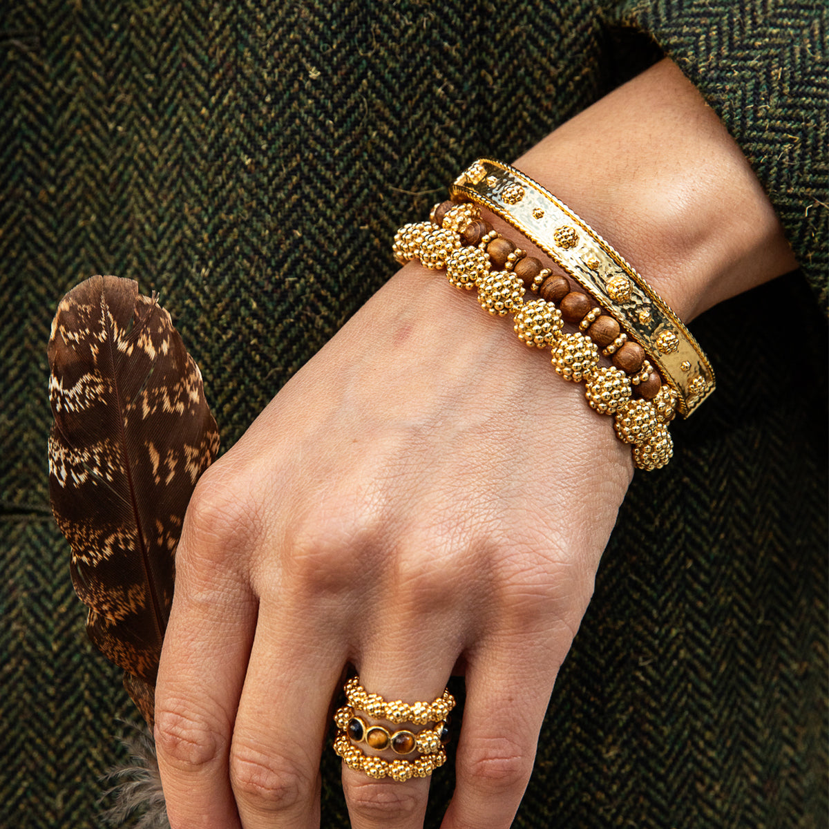 A close-up of a person's hand wearing multiple gold-toned bracelets and a matching ring, with a brown feather resting beside the hand against a dark herringbone textile background.