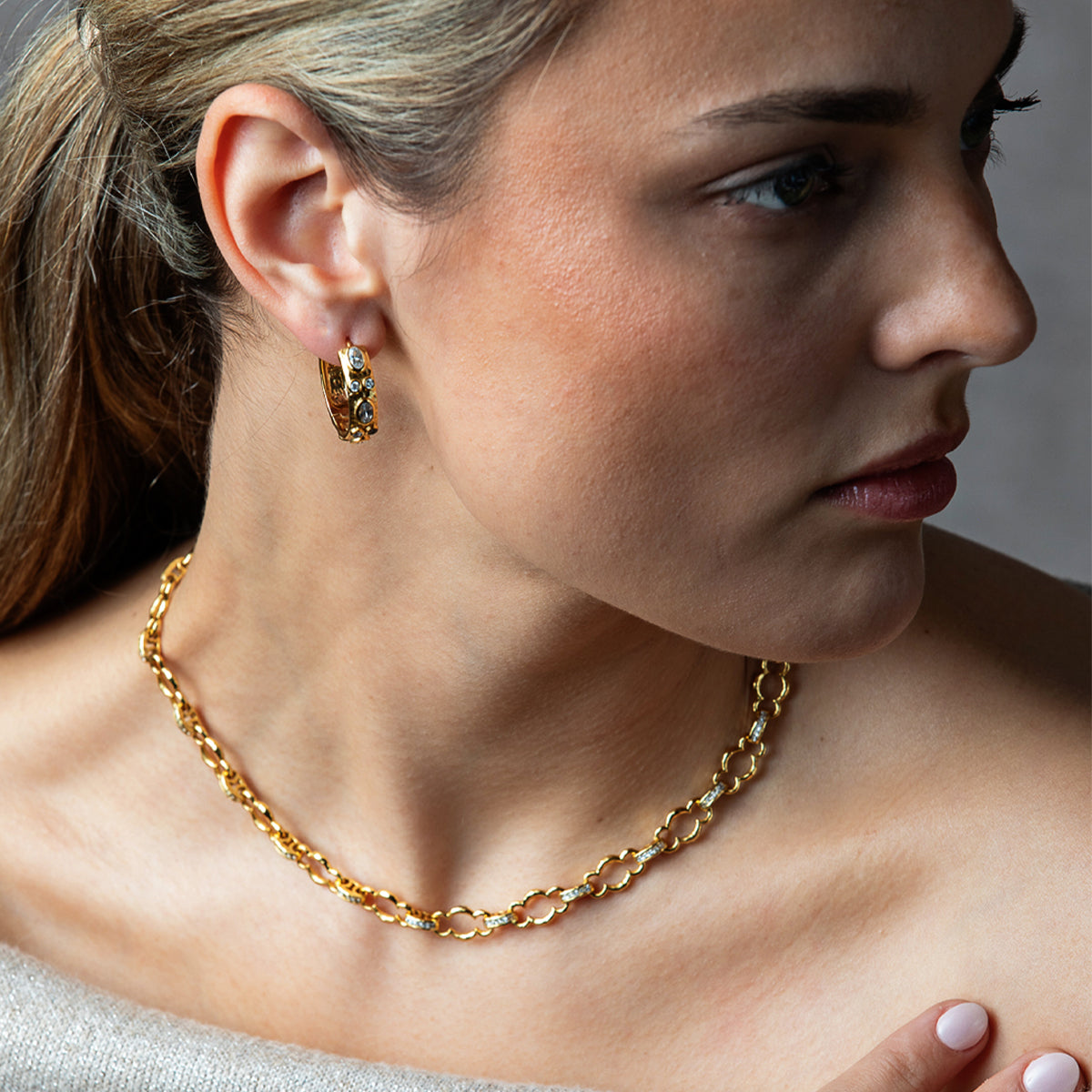 A close-up lifestyle shot of a woman wearing a delicate gold chain necklace and a matching textured gold hoop earring.