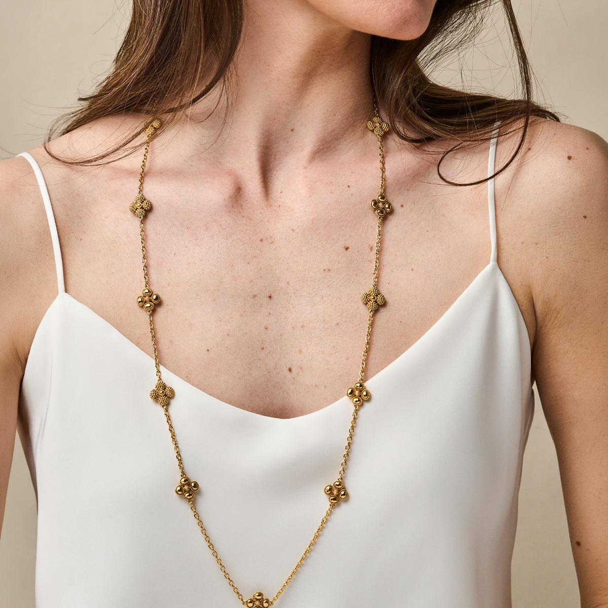 A gold chain necklace with ornate, textured floral or clover-like links, worn by a model in a white camisole top against a neutral beige background.
