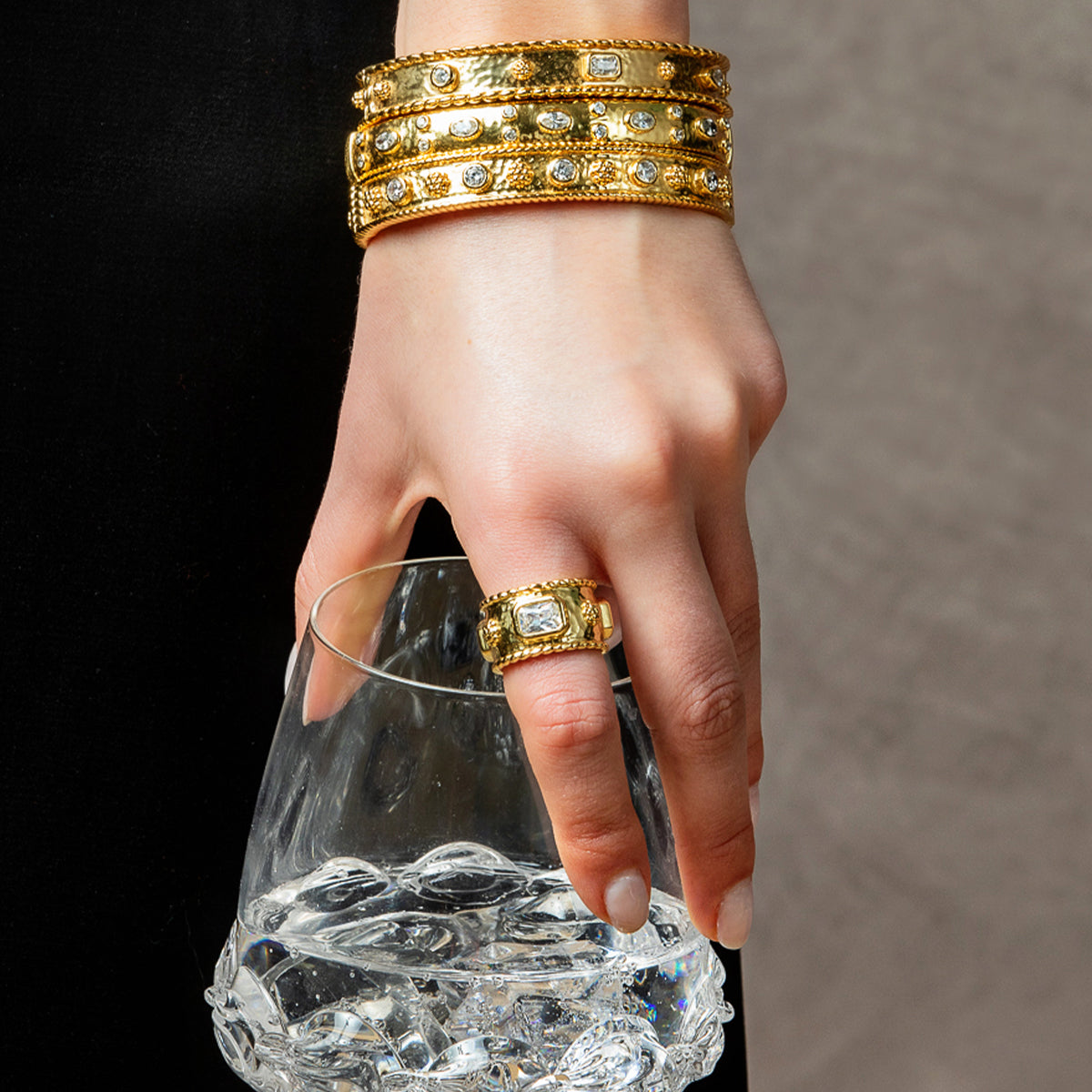 A close-up of a hand wearing a gold ring with a clear central gemstone, holding a textured glass of water. The hand also wears three stacked gold bangles with clear stones, set against a dark background.
