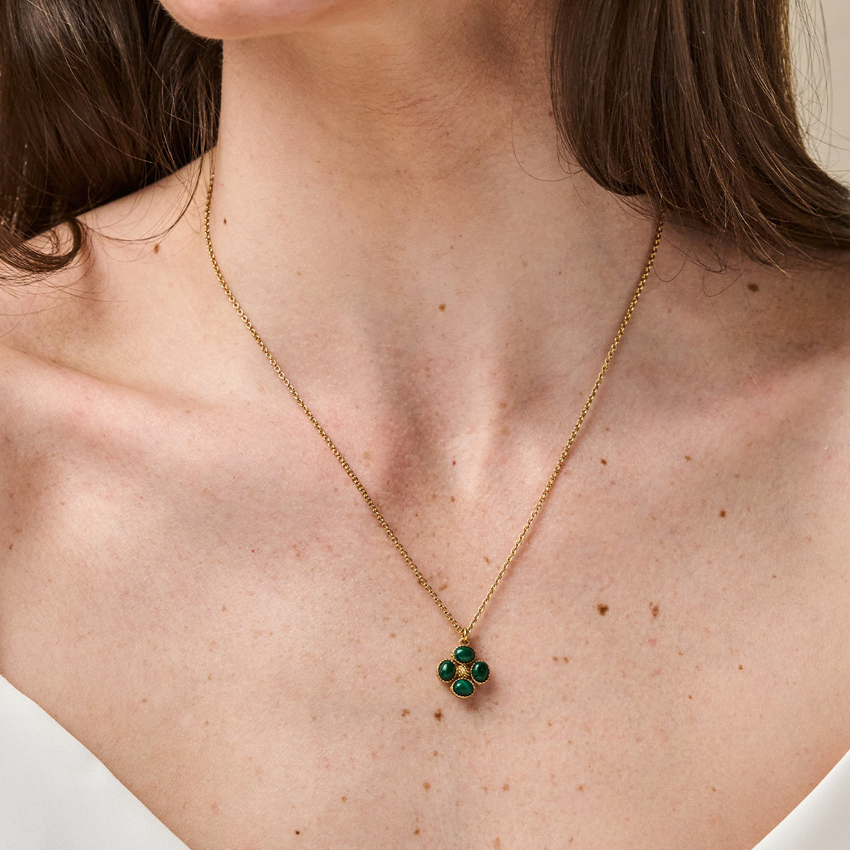 A close-up of a model's neck and upper chest wearing a delicate gold chain necklace with a small four-petal flower pendant featuring green stones.