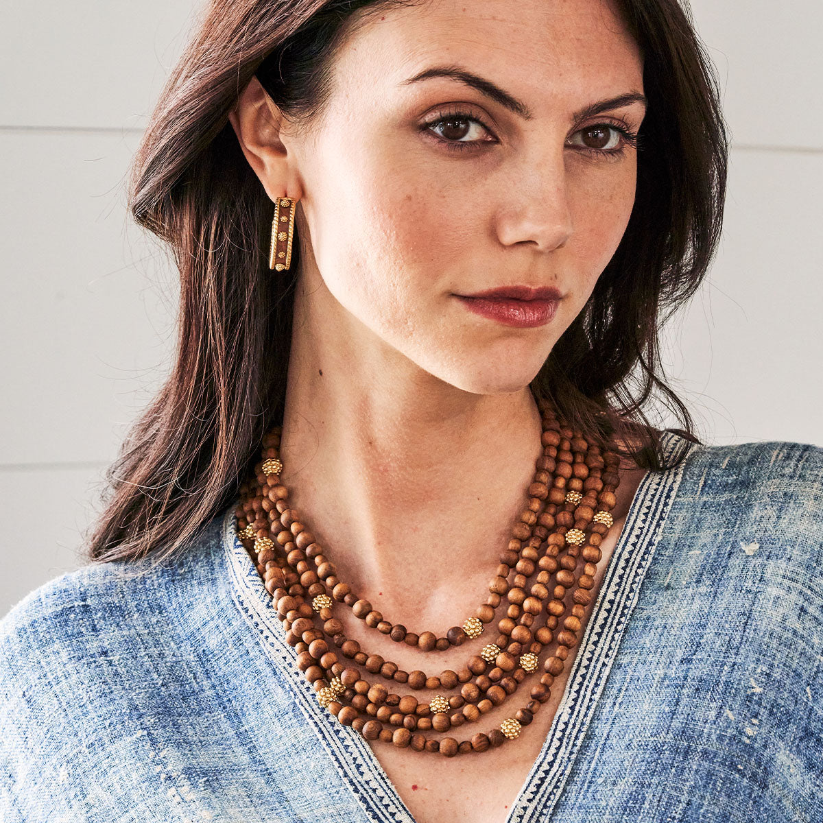 Woman wearing a stack of wooden bead necklaces and gold earrings against a neutral background