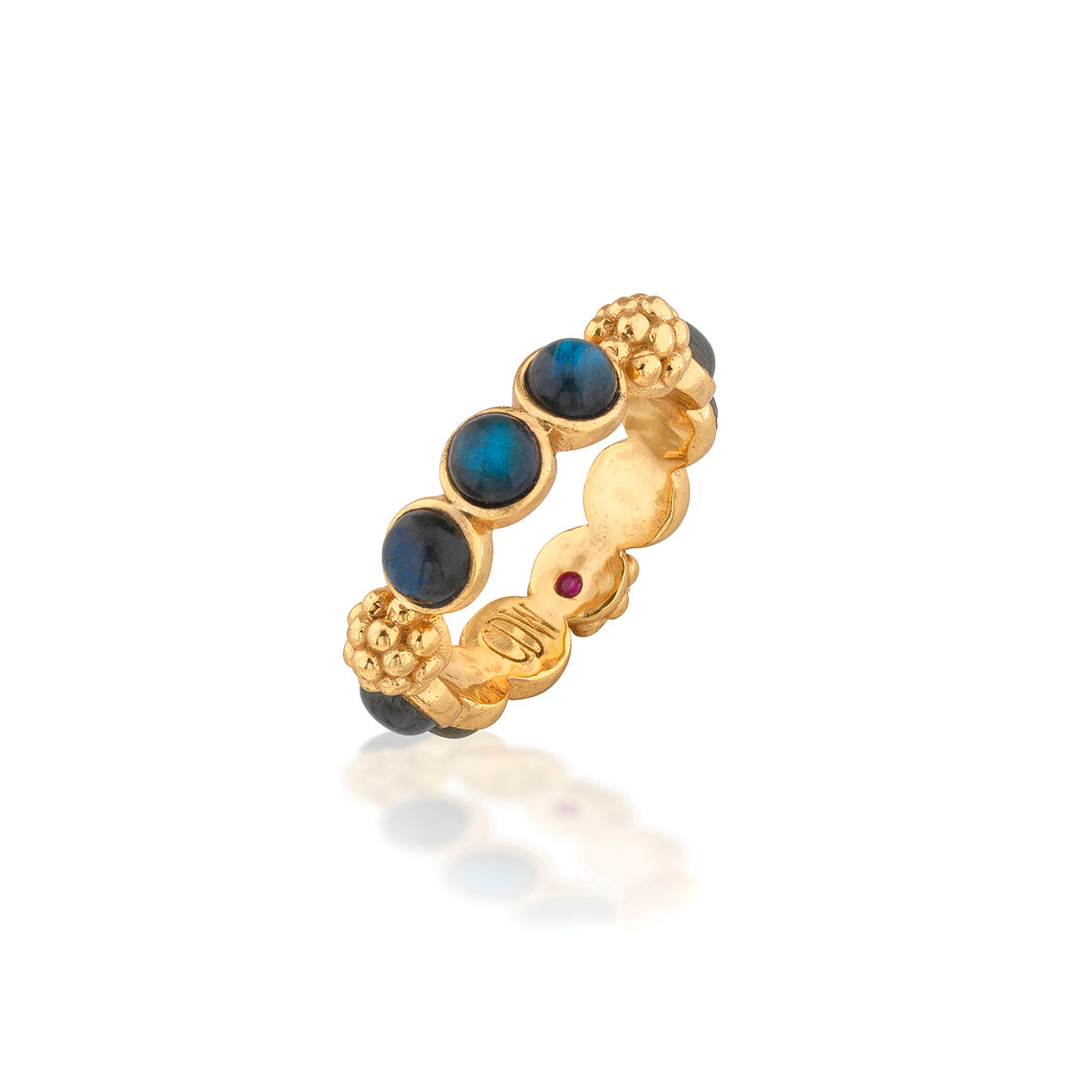 A gold band ring featuring oval dark blue cabochon stones, textured gold bead clusters, and a small round red gemstone accent, shown on a clean white background with a soft reflection.