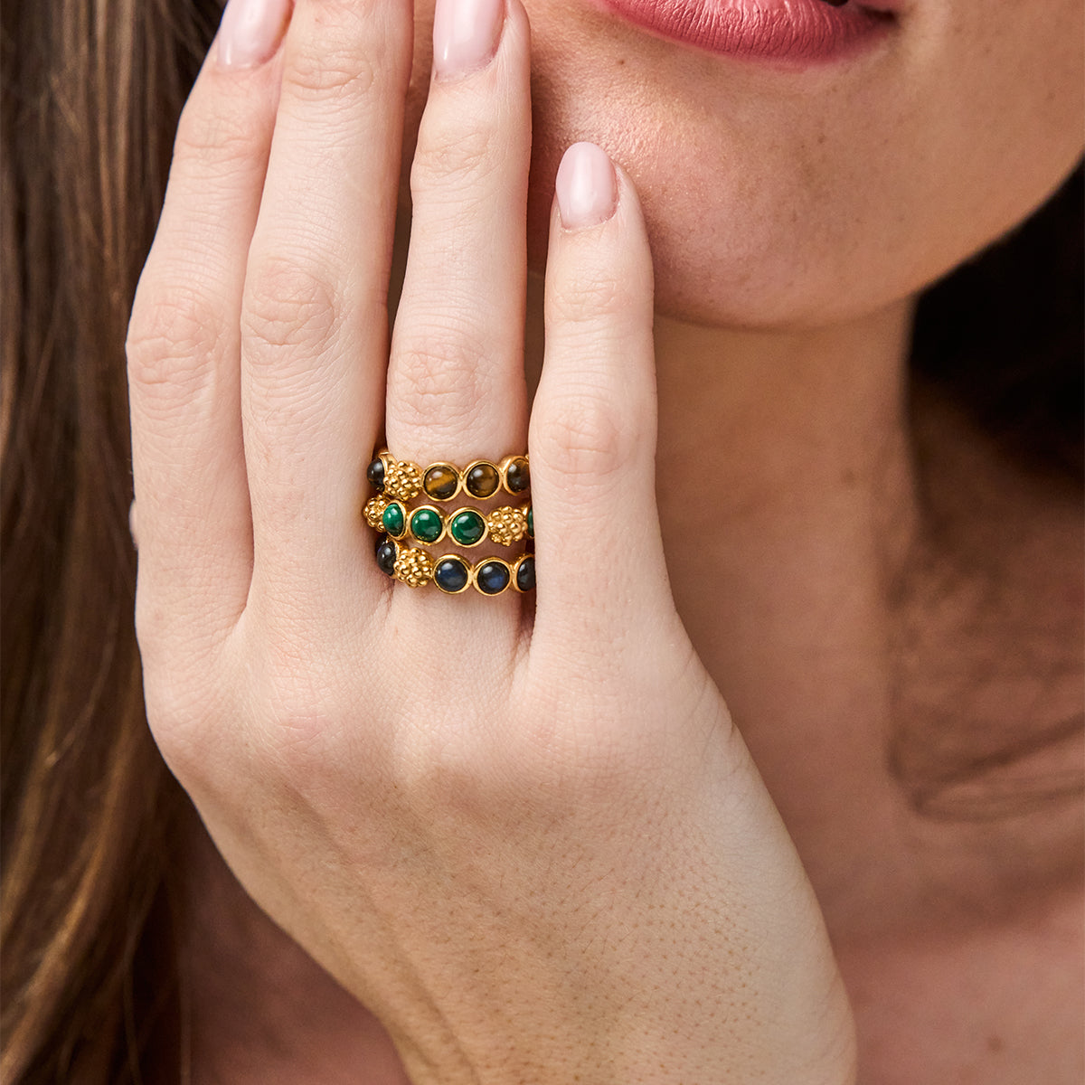 A close-up of a woman's hand wearing a stack of three gold rings decorated with green and dark blue round stones.