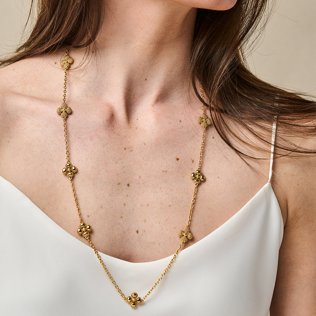 A gold necklace with textured, flower-like clover motifs draped over a woman's white camisole top.