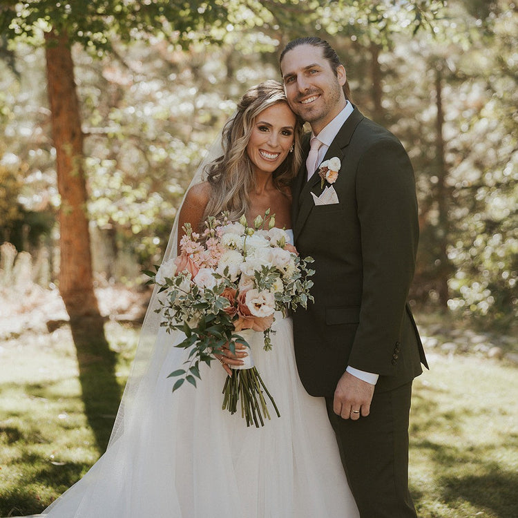 A smiling bride in a white wedding gown holding a pink and green bouquet stands next to a groom in a dark green suit outdoors under leafy trees.
