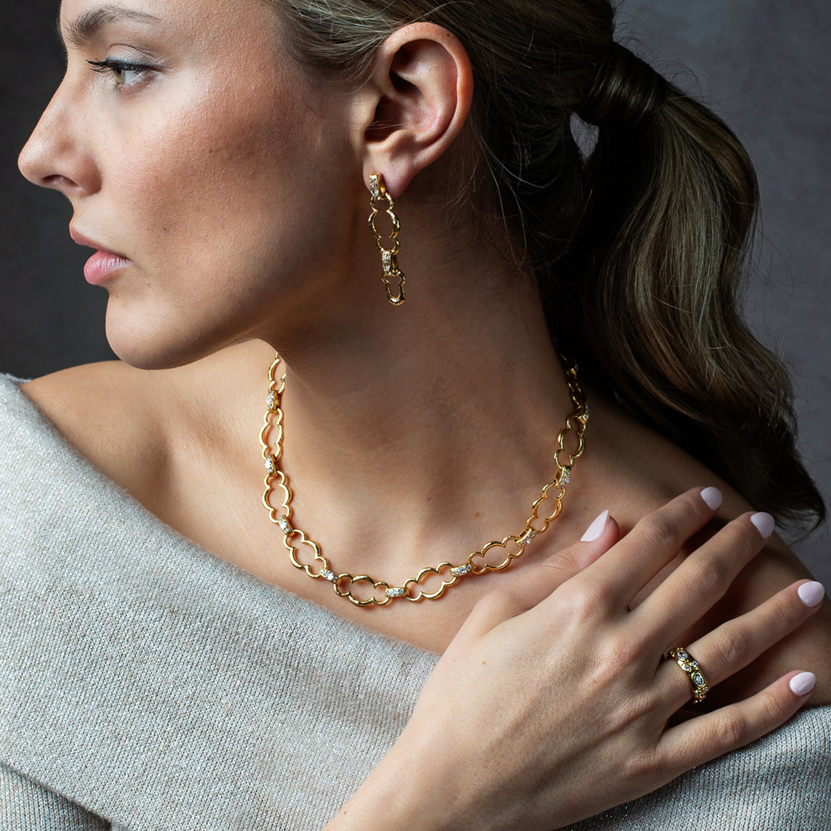 A close-up lifestyle shot of a woman wearing a gold chain necklace, matching drop earrings, and a ring, styled with a gray knit top against a dark gray background.