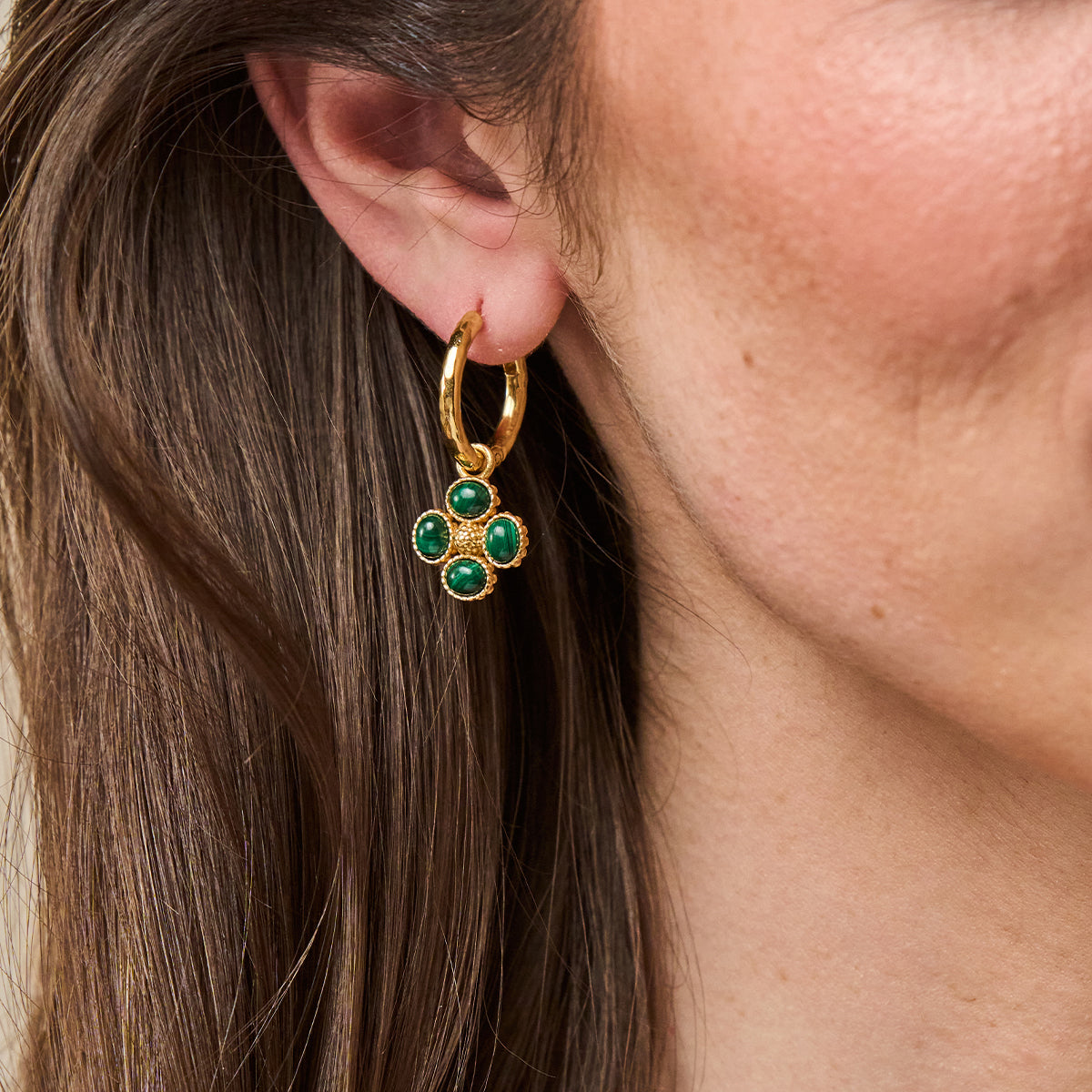 A close-up of a model's ear wearing a gold hoop earring with a dangling four-petal flower charm set with green stones.