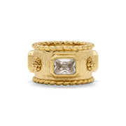 A wide yellow gold ring with a textured hammered center, a rectangular clear gemstone, and decorative grape motifs on the sides, framed by rope-textured borders on a white background.