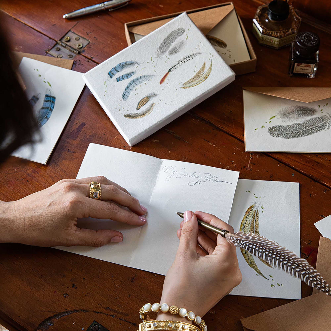 Person writing with a feather pen on a wooden desk with stationery items.
