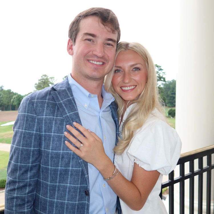 A smiling couple posing together on a porch. The man wears a blue plaid blazer over a light blue button-down shirt and khaki pants, while the woman wears a white puff-sleeve top.