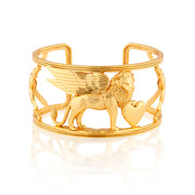 A gold-tone cuff bracelet featuring a winged lion holding a heart, with decorative chain-like side details, shown on a clean white background with a soft reflection below.