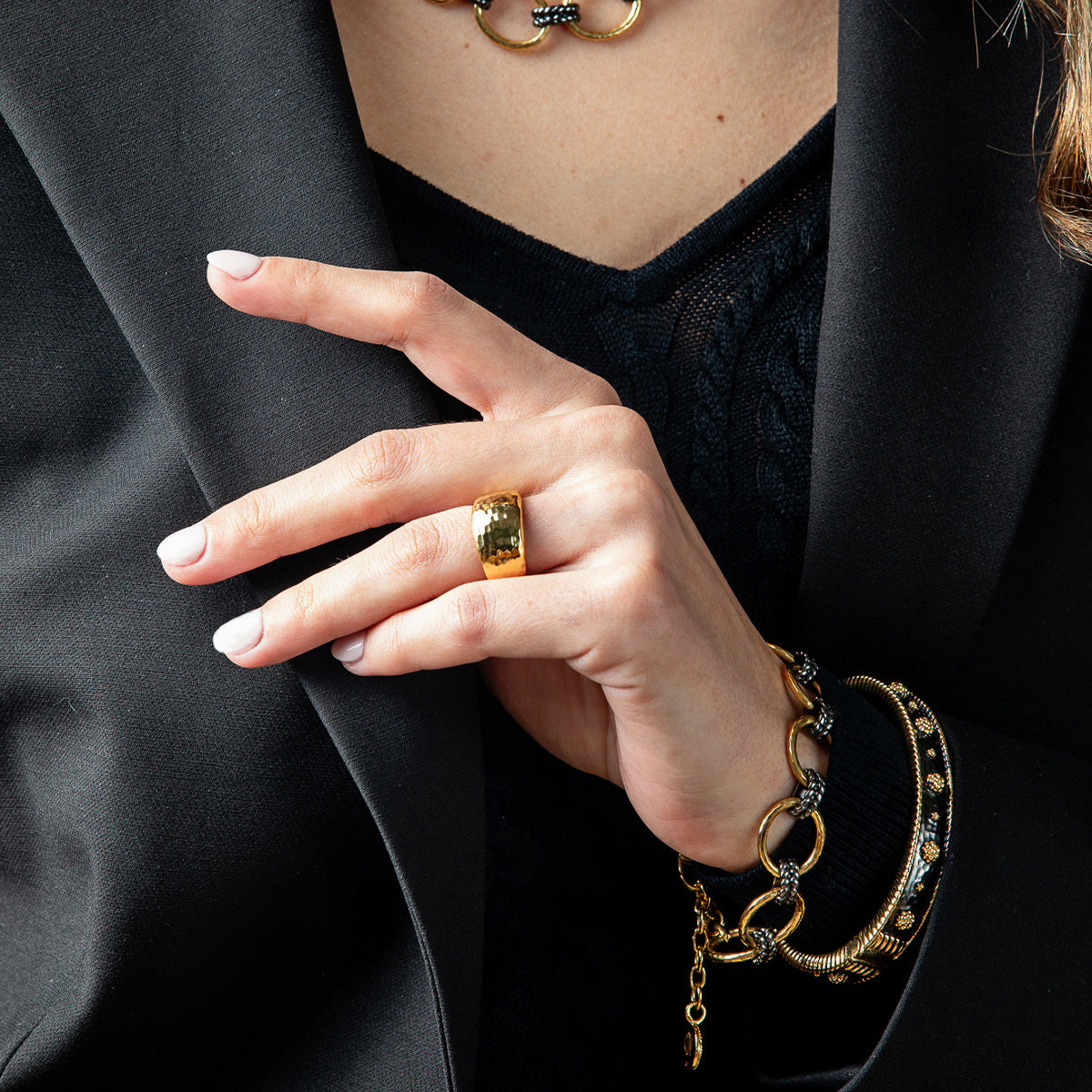 A close-up of a person's hand wearing a chunky gold ring, styled with a black blazer, black top, and gold chain and bangle bracelets.