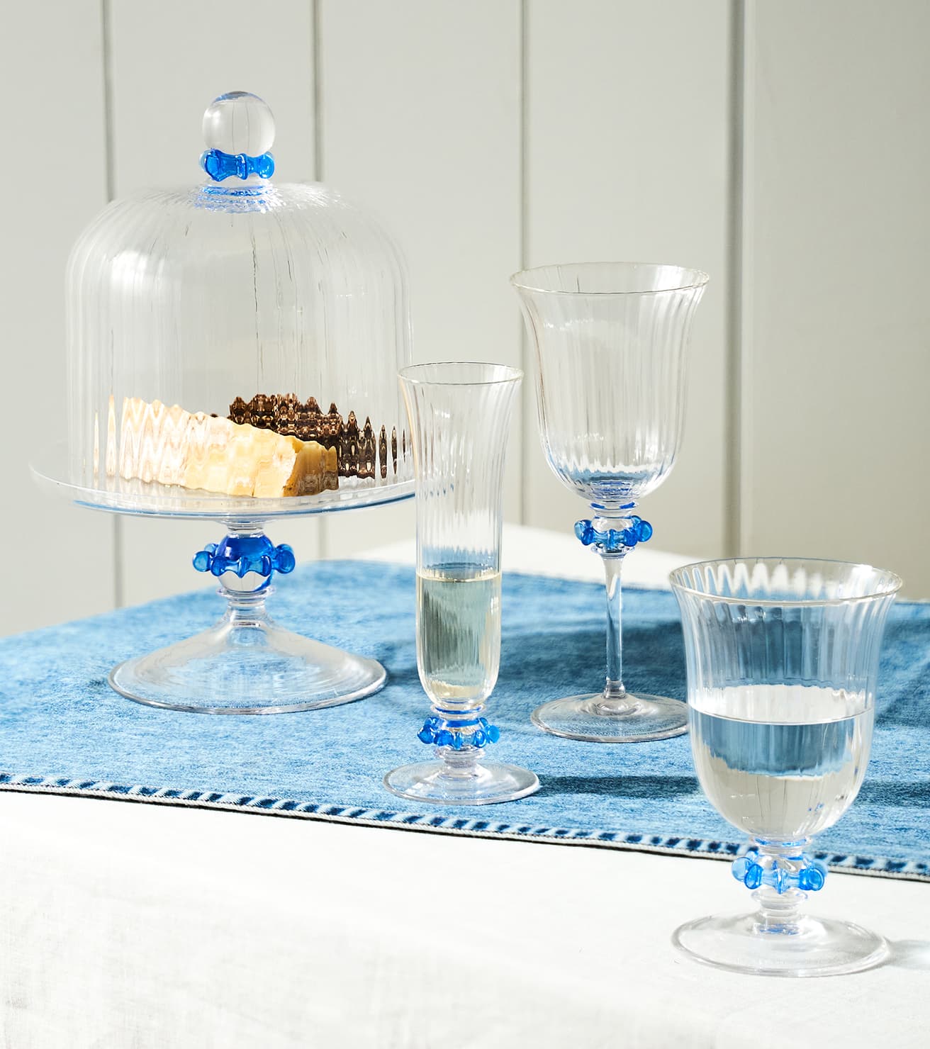 A set of clear glass tableware with bright blue decorative accents, including a cake dome with food, a flute glass, a goblet, and a tumbler, arranged on a blue runner over a white table.