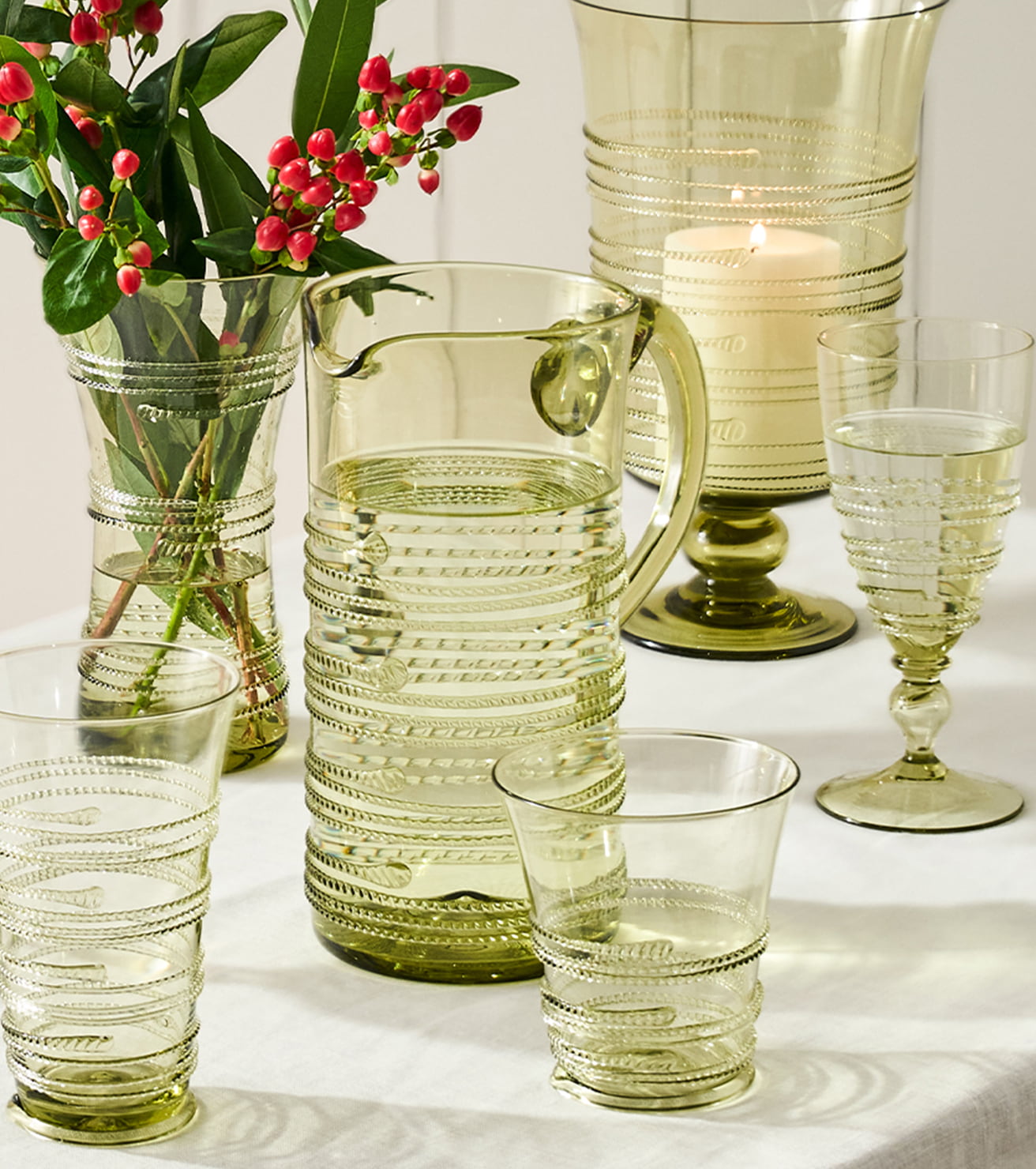 A set of ribbed clear glass tableware in a light olive-green tint, including a pitcher, tumblers, a goblet, and a large vase, arranged on a white tablecloth with a lit candle and red berries in the background.