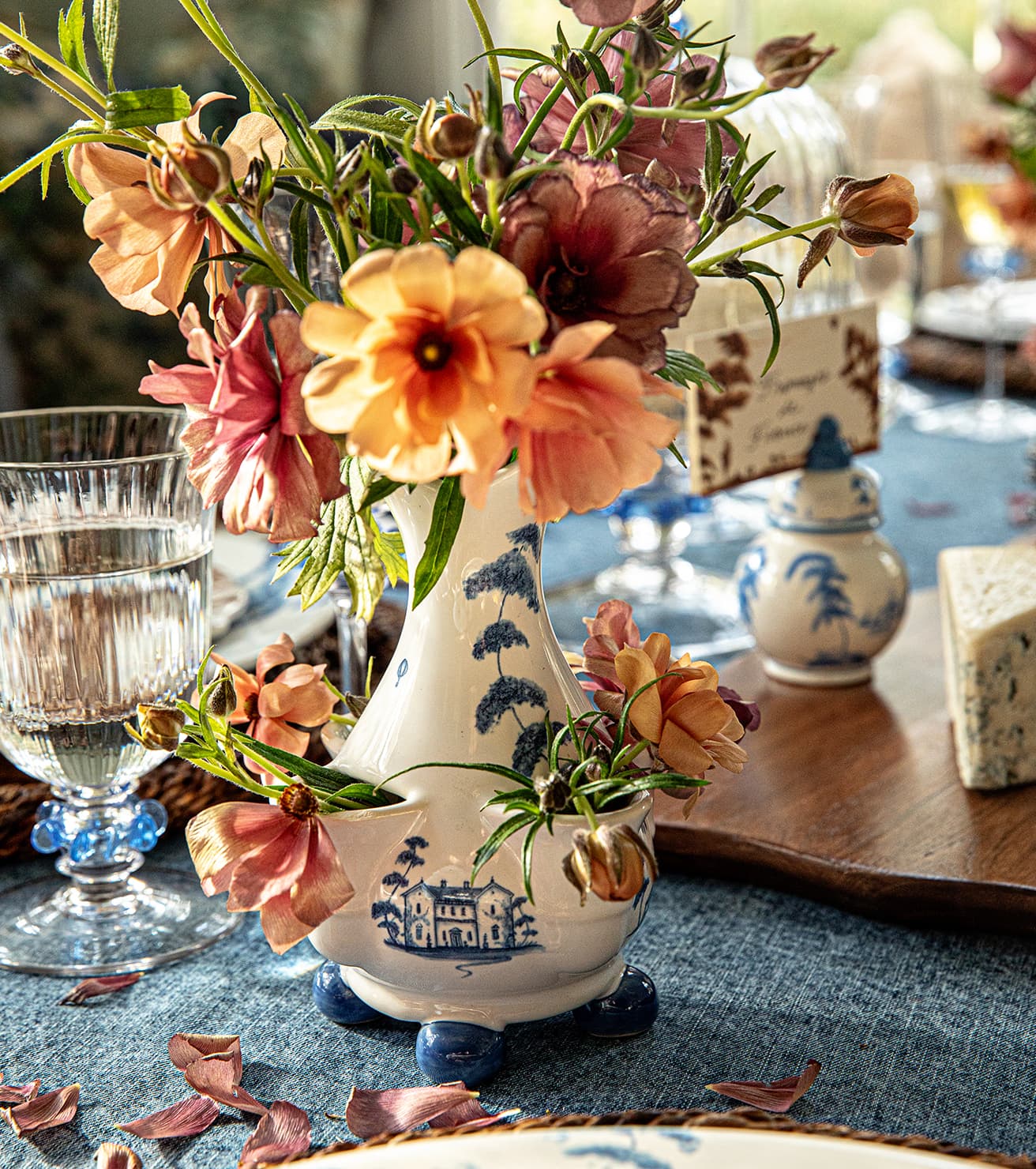 A decorative ceramic vase filled with orange, pink, and peach flowers, styled on a blue tablecloth with a glass, blue goblet, and cheese in a dining setting.