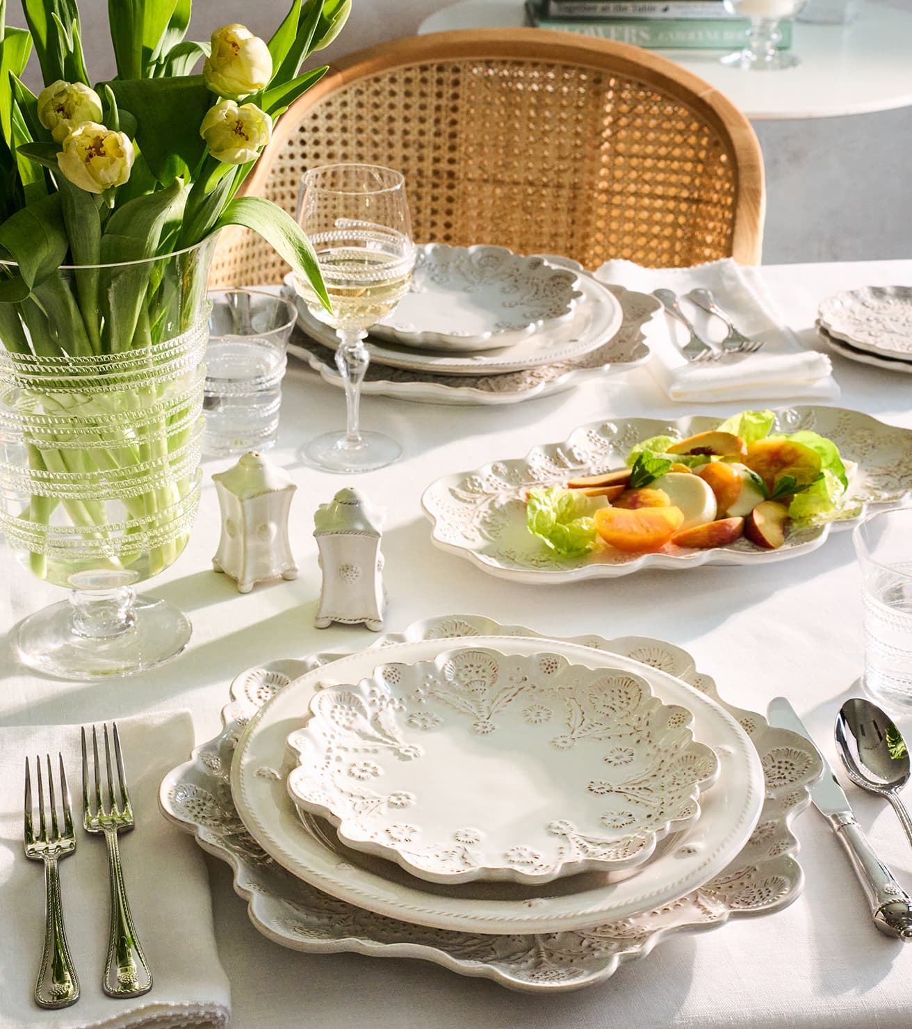 A beautifully styled dining table setting featuring a ceramic dinnerware set with intricate floral cutout detailing. The set includes scalloped plates, matching bowls, and silver cutlery on a white tablecloth, accompanied by a glass vase of fresh tulips and a woven cane chair in the background.