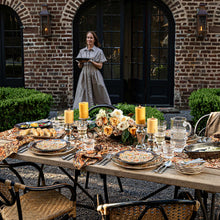 Woman standing behind a beautifully set outdoor dining table with candles and flowers, against a brick wall.