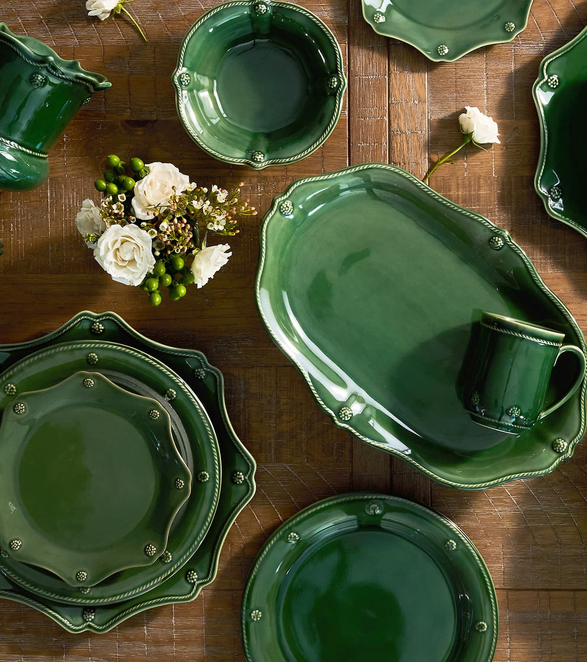 A coordinated set of glossy dark green ceramic tableware arranged on a wooden table, including plates, bowls, a serving tray, a mug, and a pitcher, styled with white flowers and greenery.
