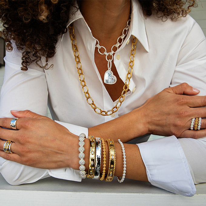 A close-up of a person wearing multiple layered necklaces and bracelets, including a chunky gold chain with a silver heart pendant, several gold and silver bangles, and pearl-style bracelets, paired with a white collared shirt.