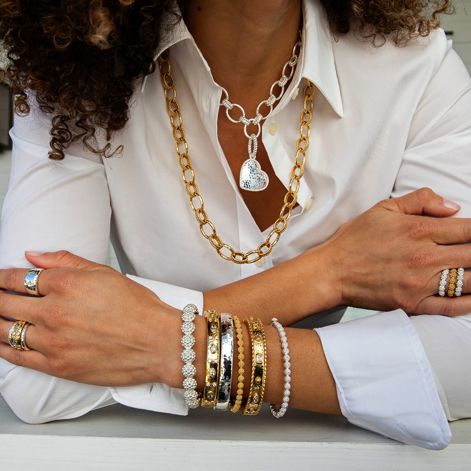 A close-up lifestyle shot of a person wearing multiple styles of jewelry, including layered necklaces (a gold chain with a silver heart pendant and a silver link chain), several gold and silver bangles on the wrist, and rings on the fingers, all styled with a white button-down shirt.