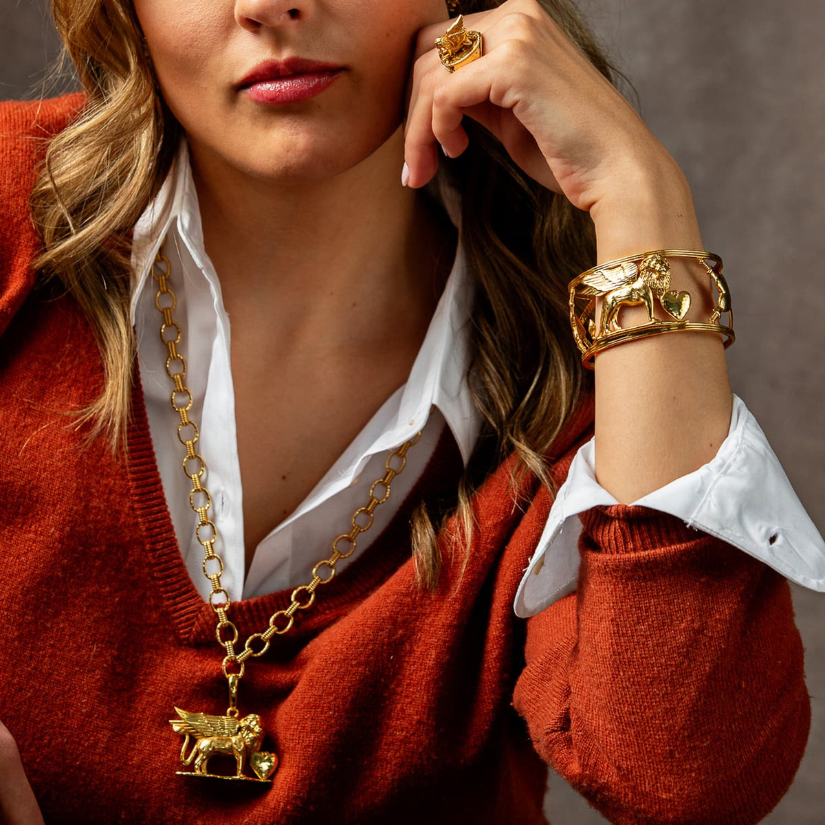 A close-up lifestyle shot of a model wearing a gold animal-themed pendant necklace and a matching gold cuff bracelet, styled with a white collared shirt and a rust-orange knit sweater against a neutral gray background.
