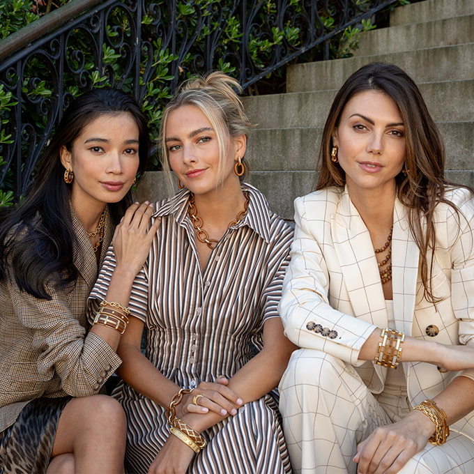 Three women sitting together on a staircase wearing stylish outfits and accessories.