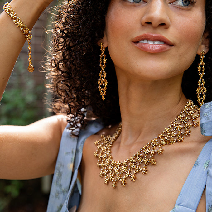 Woman wearing gold jewelry including a necklace, earrings, and bracelet outdoors.