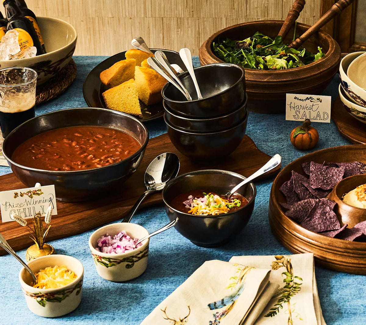 A styled autumnal dining table featuring a variety of hearty dishes in rustic ceramic and wooden bowls, including a large bowl of chili, a bowl of salad, cornbread, and tortilla chips, set against a blue tablecloth.
