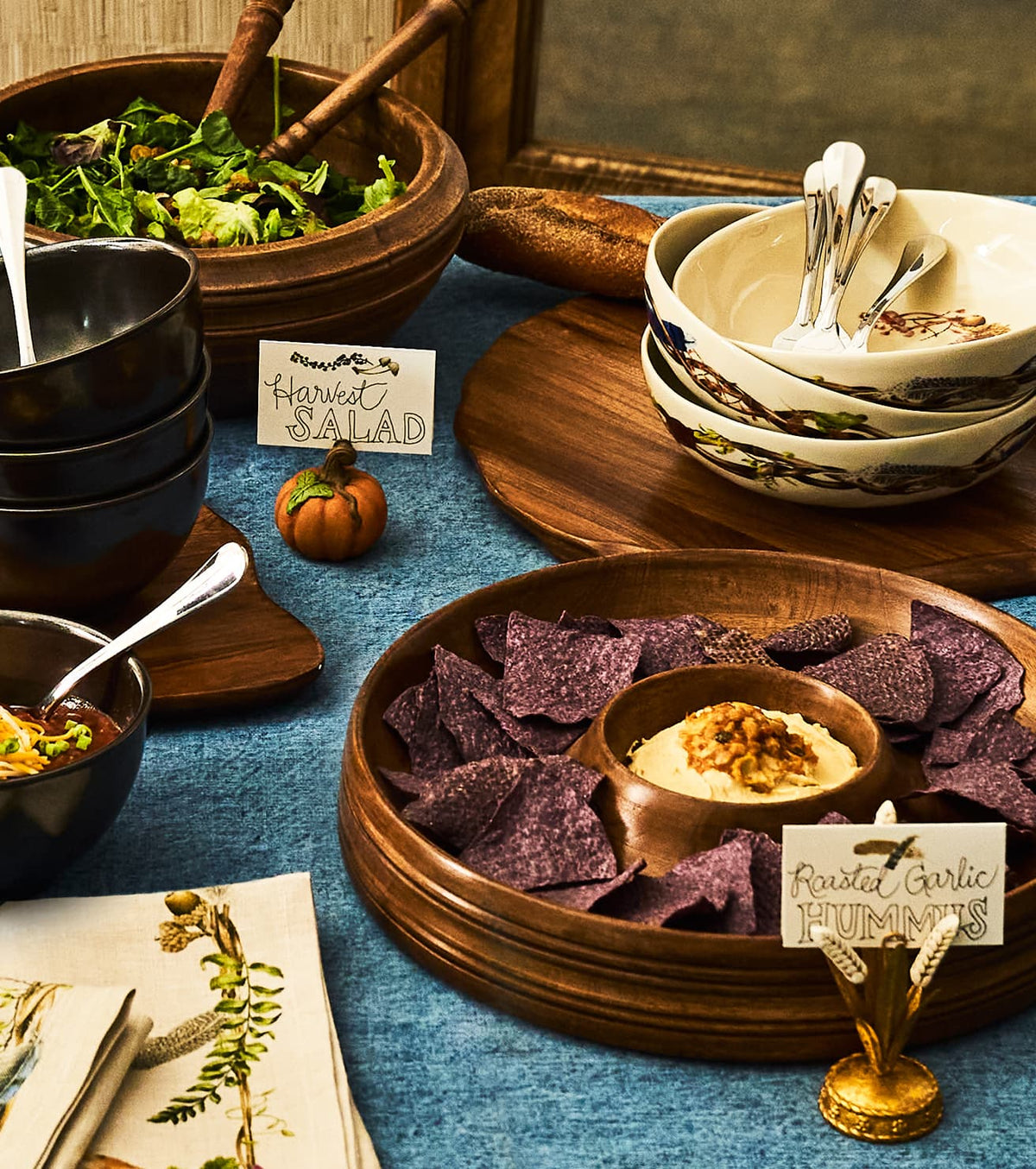 A styled food table setting with multiple bowls and wooden serving trays. In the foreground, a round wooden tray holds purple tortilla chips and a small bowl of creamy dip with a small candle.