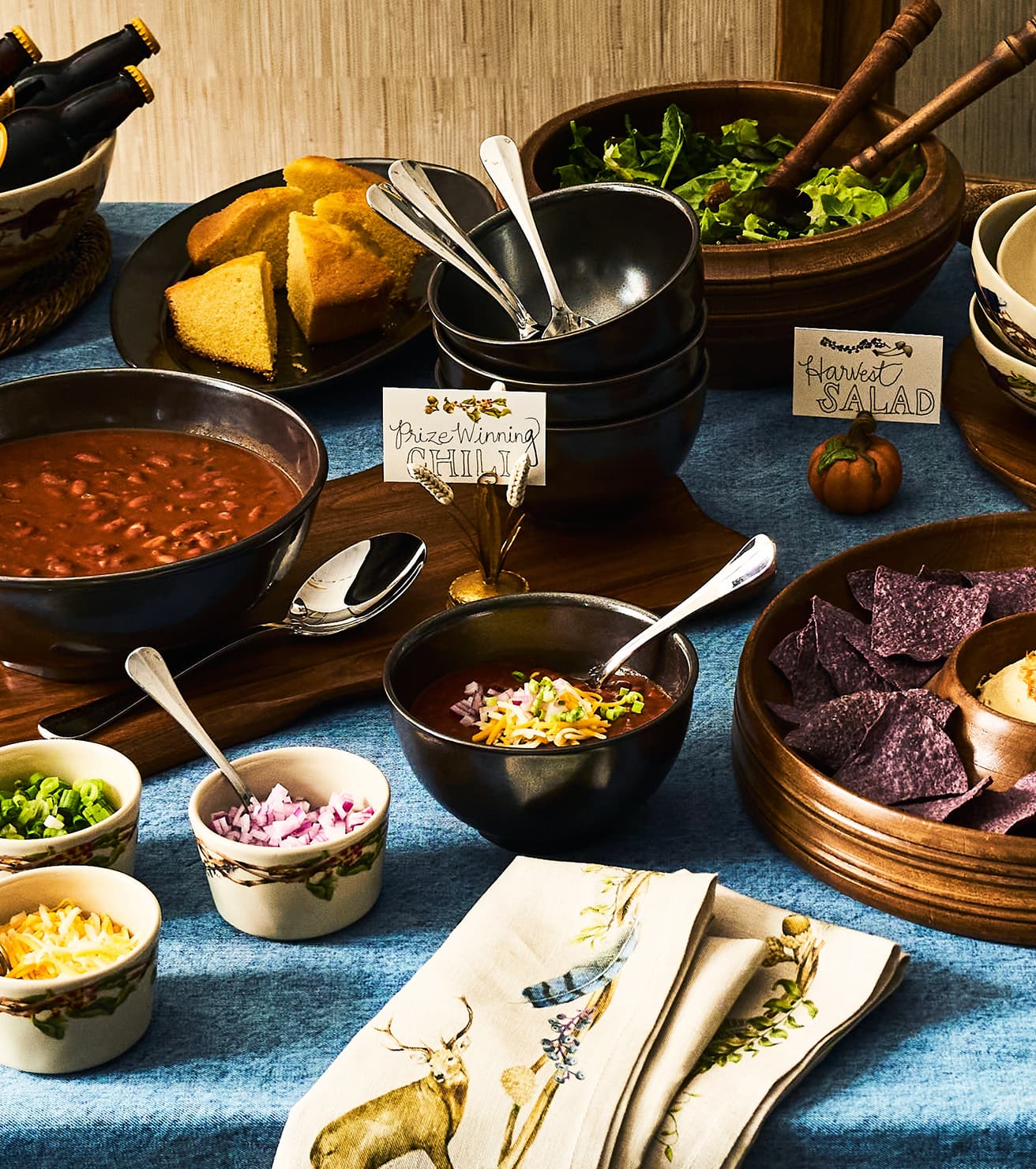 A styled food spread on a blue tablecloth with black bowls of chili and soup, a plate of cornbread, small bowls of chopped onions and cheese, purple tortilla chips, and a folded cream napkin with a deer print. Handwritten signs read 'Prize Winning Chili' and 'Harvest Salad'.