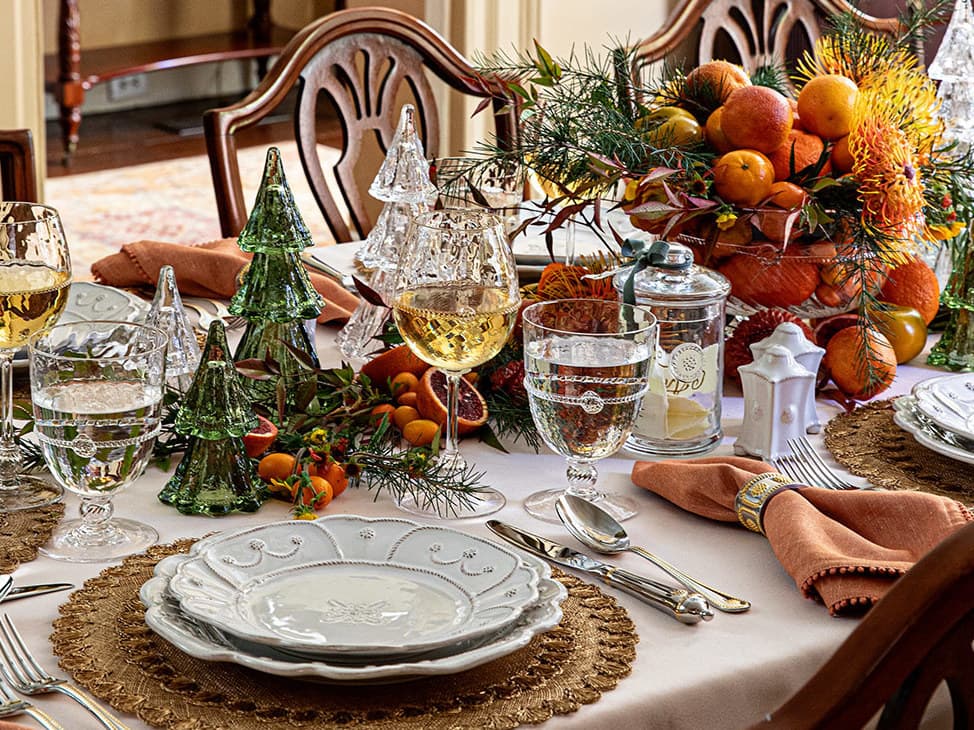 A beautifully set dining table for a holiday gathering, featuring white patterned plates on woven placemats, clear glassware, orange and gold floral centerpieces, and folded orange napkins.