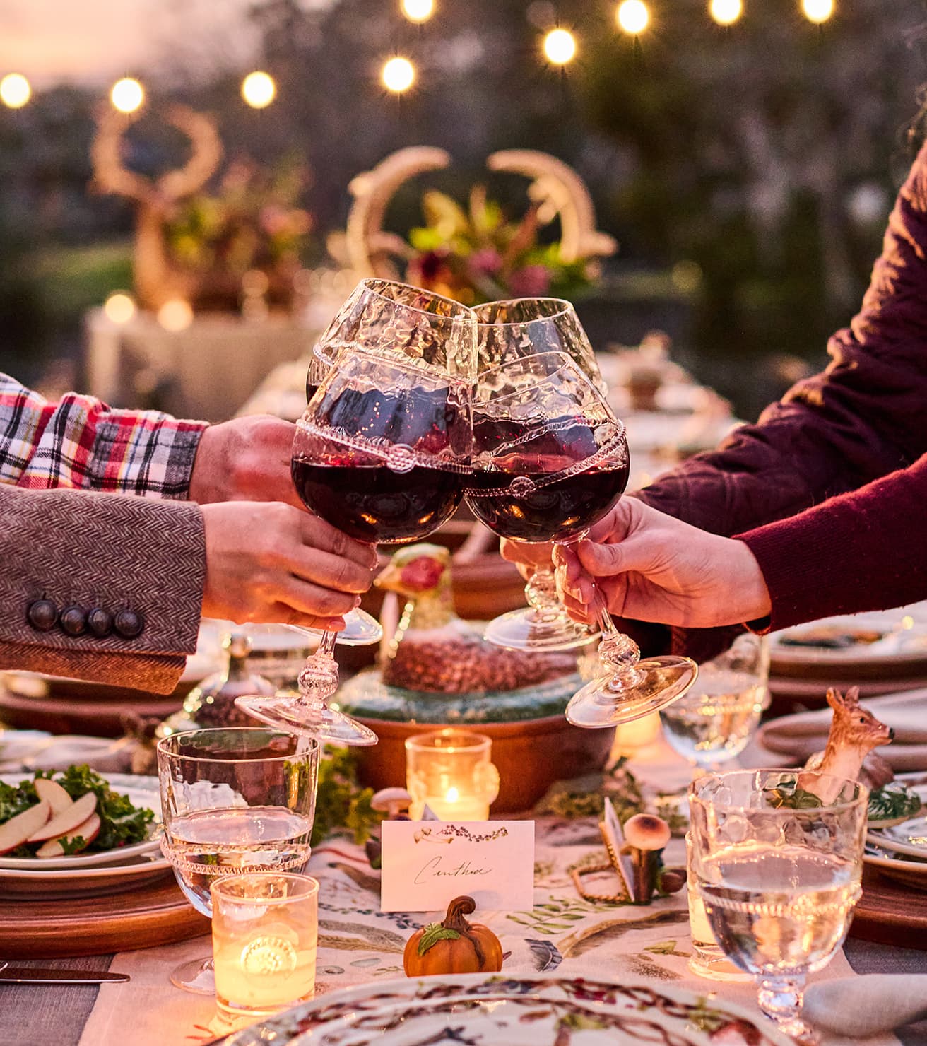 Two people clinking wine glasses at a festive outdoor dinner table with string lights and autumn decorations.